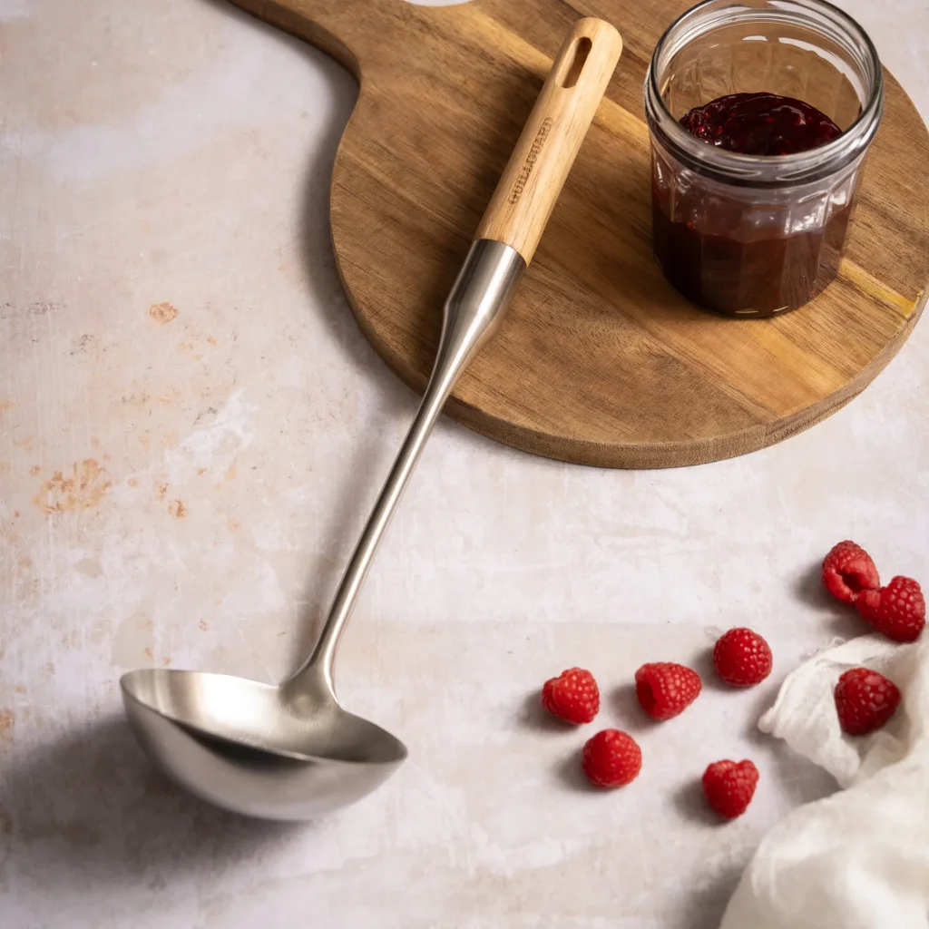 Louche en inox avec manche en bois posée à côté d’un pot de confiture et de framboises fraîches
